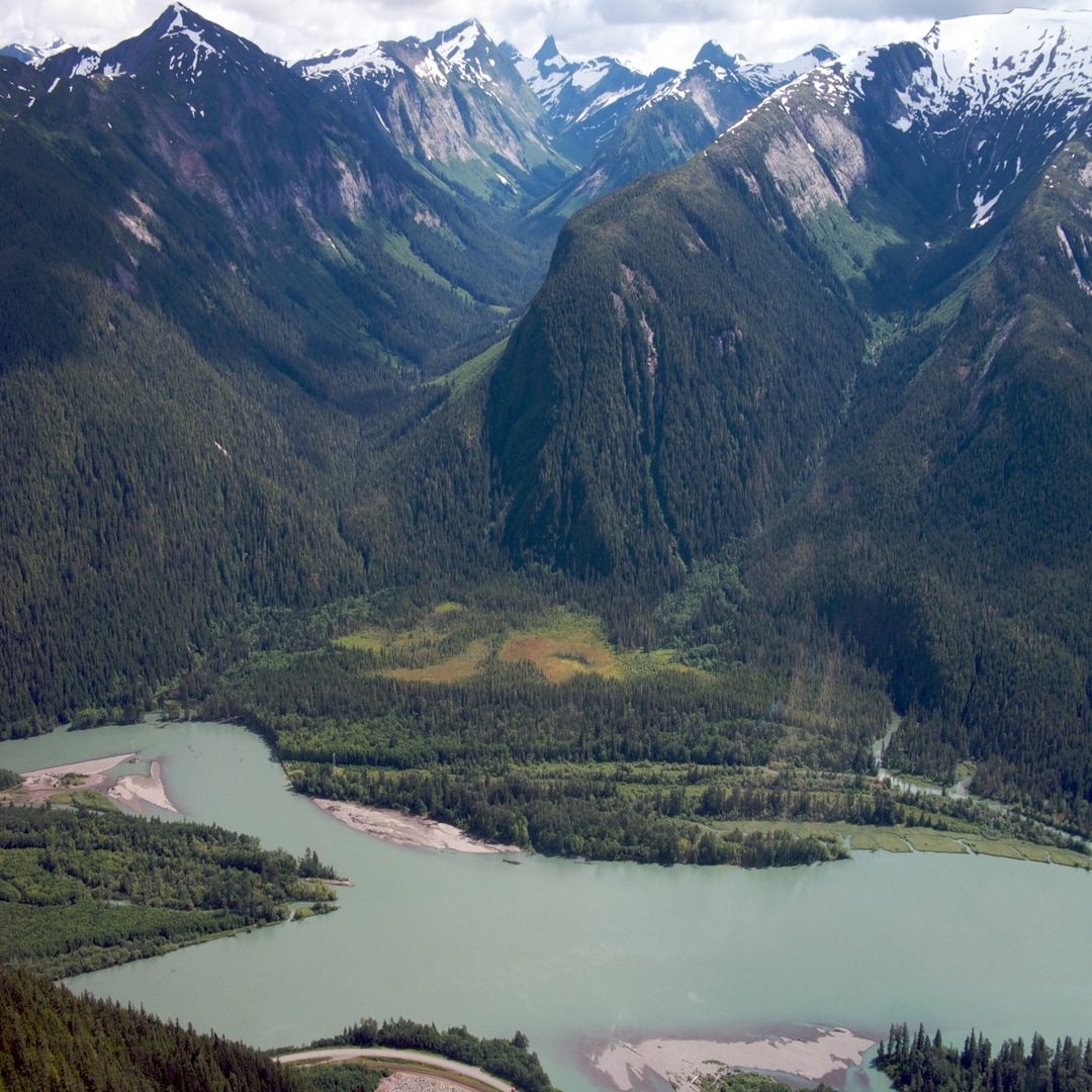 Skeena_Riding_Association A panoramic view of the Saanich South region from a rocky hilltop, showcasing lush green fields, farmland, and suburban areas under a cloudy sky.